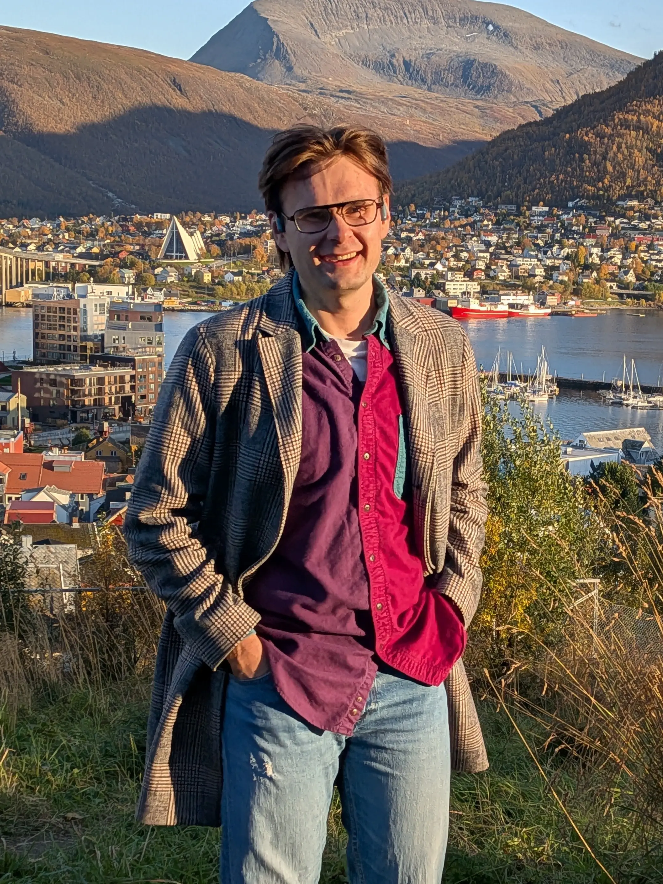 Man wearing glasses and a plaid coat standing on a hillside with a scenic view of a waterfront town and mountains in the background.