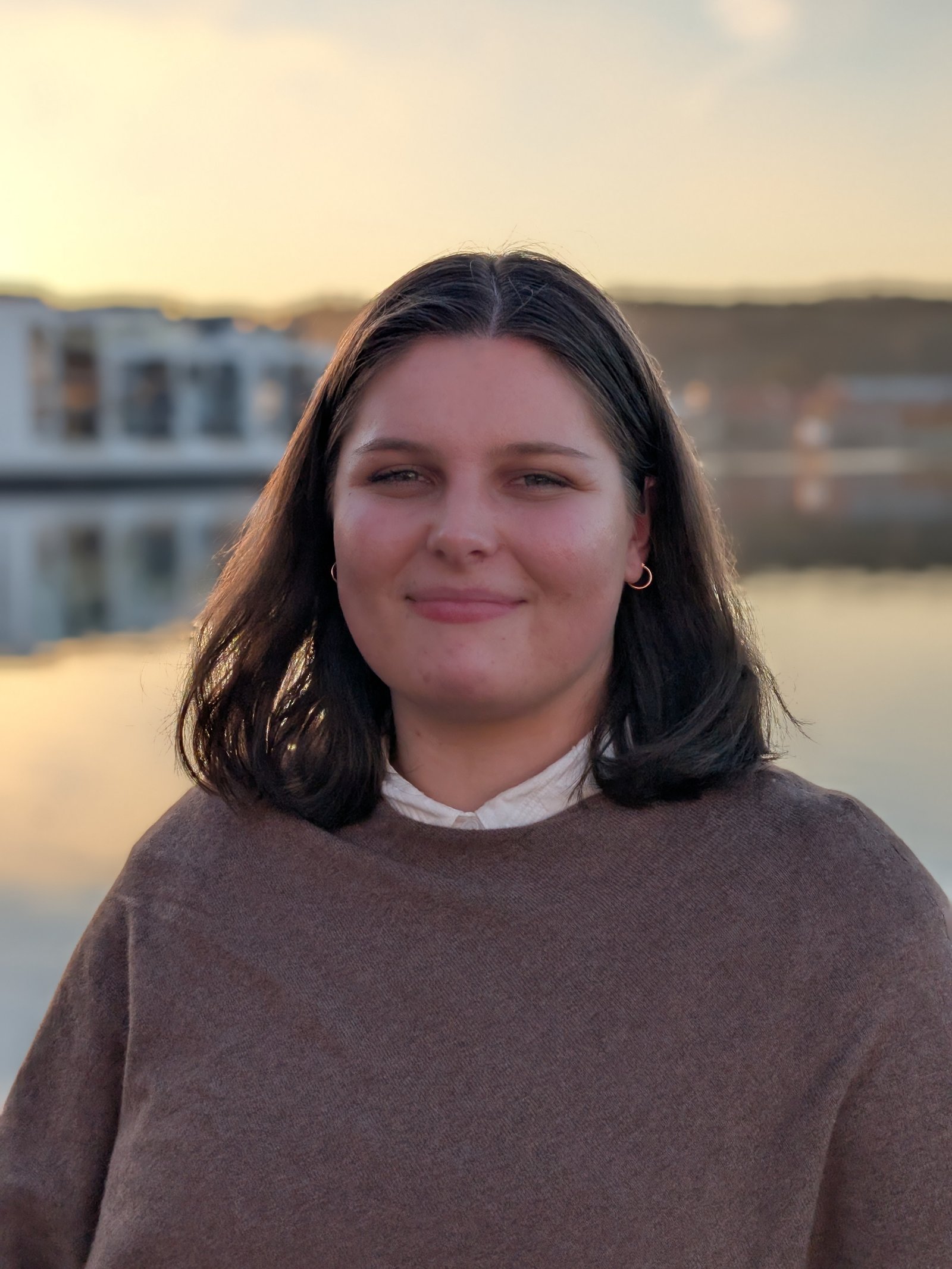 Young woman with shoulder-length dark hair, wearing a brown sweater, standing outdoors by calm water at sunset.